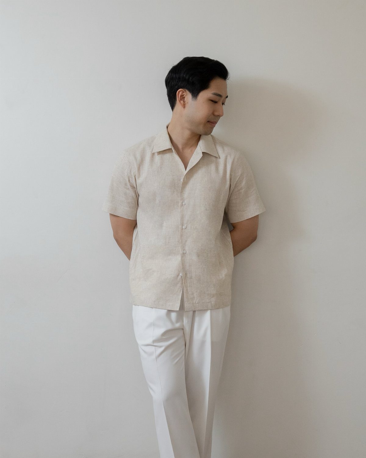 Man in a beige short-sleeve linen button-down shirt and white pants leaning against a wall.