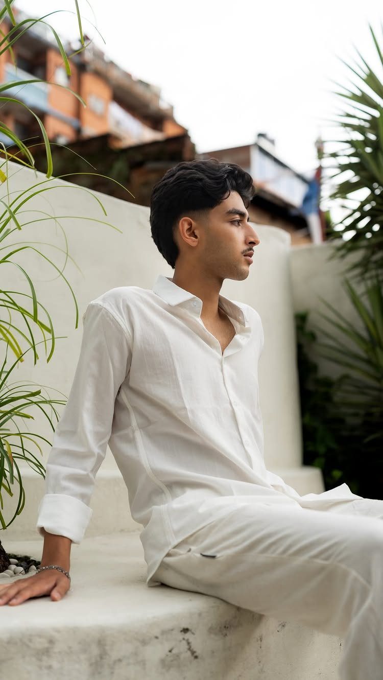 A man wearing a white linen long-sleeve shirt and matching linen pants sitting on white steps.