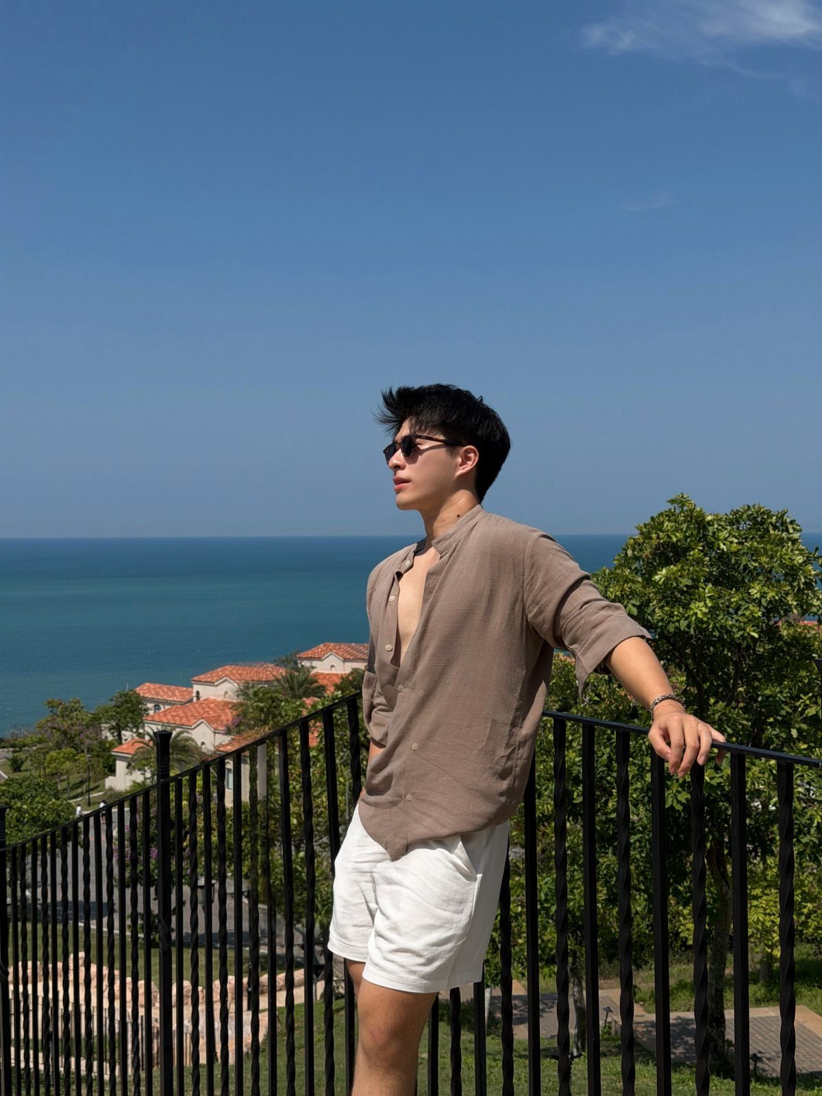 Man in a brown shirt and white shorts standing by a railing overlooking the ocean.