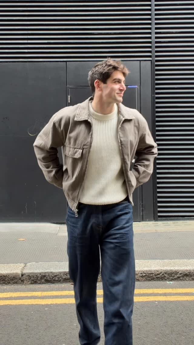 A man wearing a cream sweater under a tan jacket and dark blue trousers.