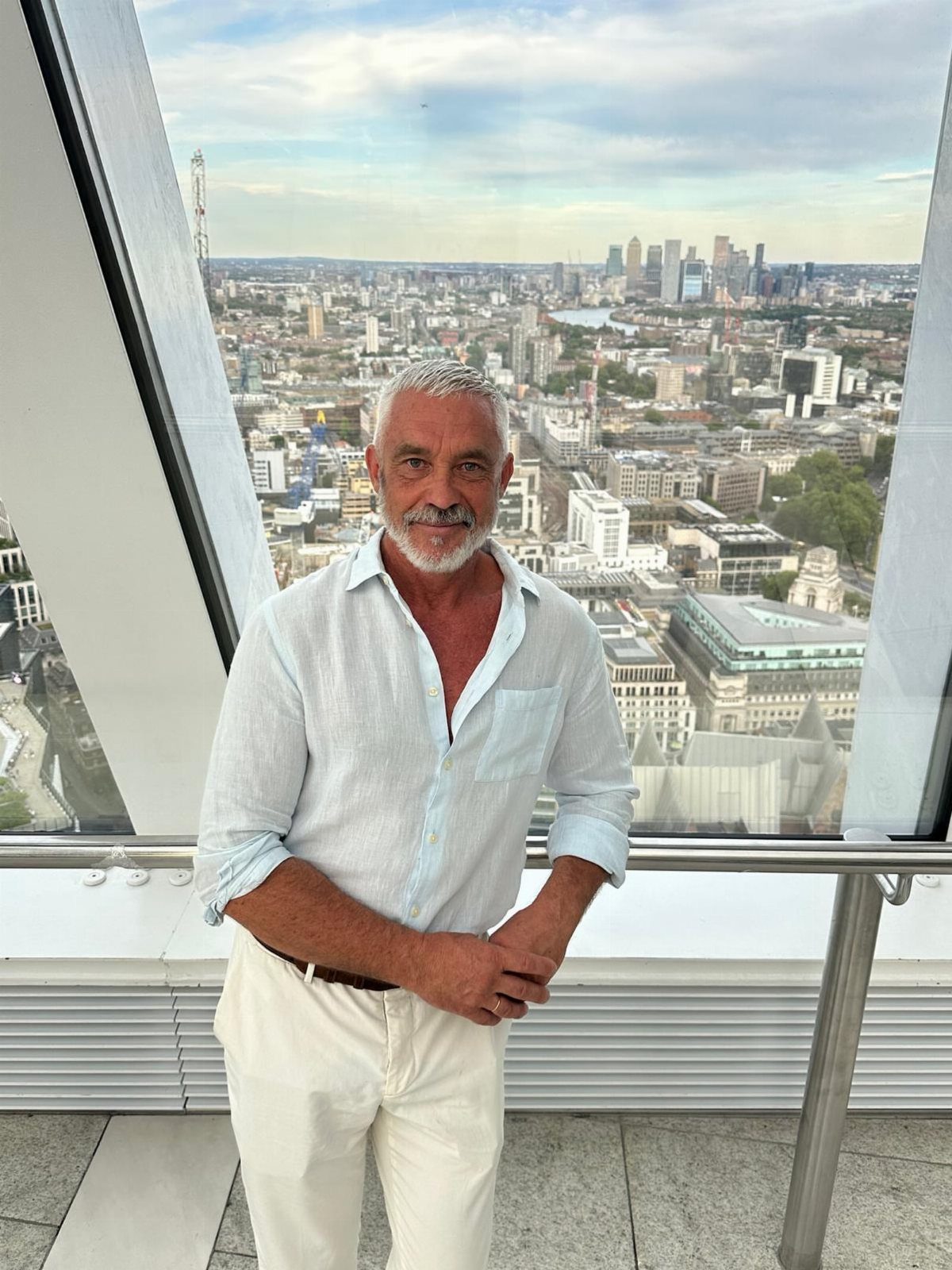 A man in a light blue linen shirt and white trousers standing against a London cityscape.