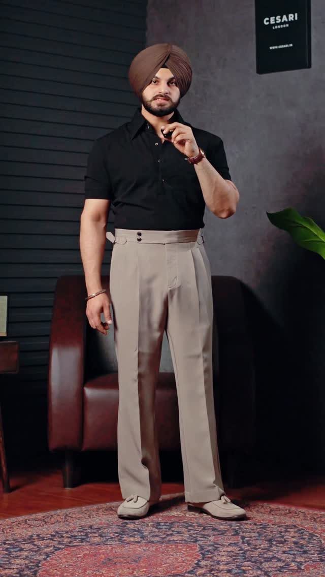 A man posing in a black shirt and beige pleated lightweight trousers.
