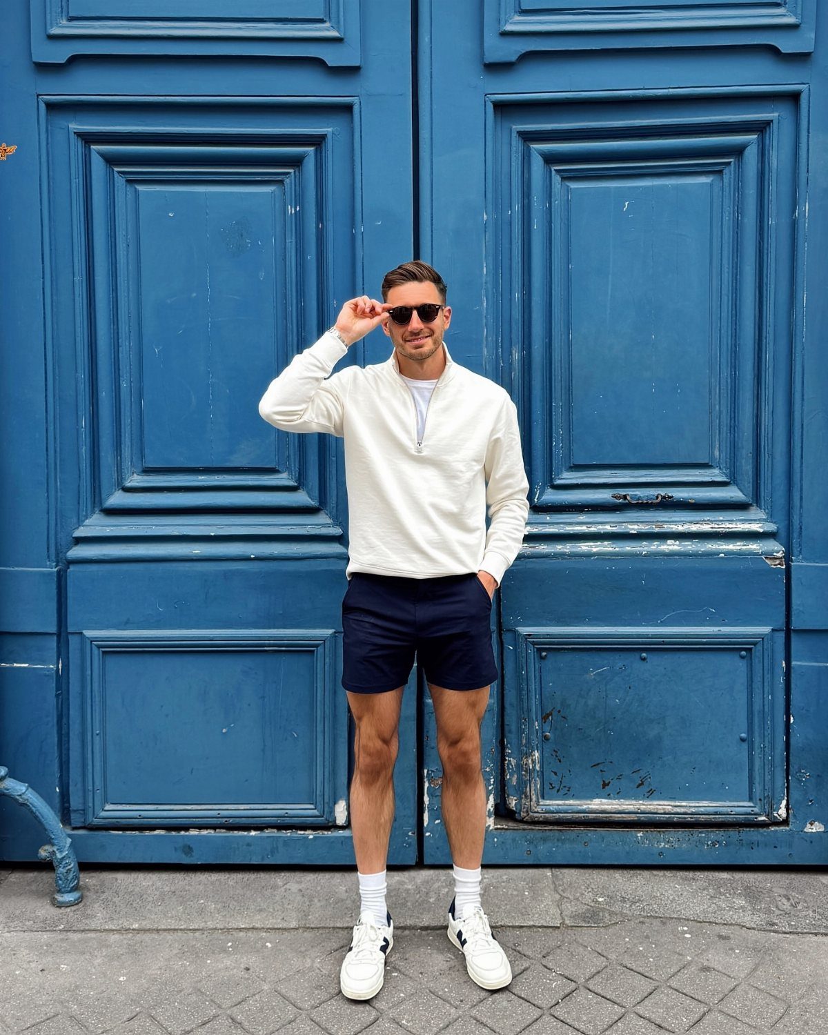 Man wearing a white pullover, navy shorts, white socks, and sneakers standing before a blue door.