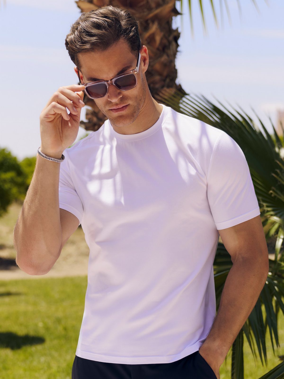 A man in a white well-fitted T-shirt and sunglasses standing outdoors near palm trees.