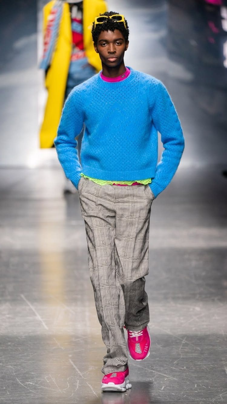 Male model walking a runway in a blue sweater, patterned trousers, and pink sneakers.