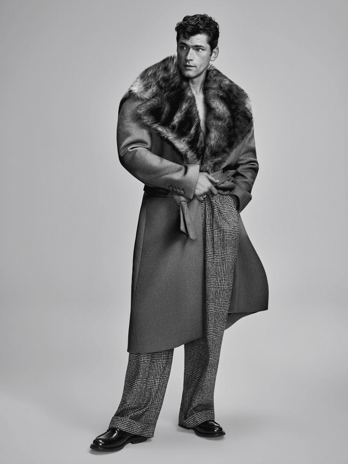 A male model wearing a fur-collared coat and patterned trousers posing in a studio.