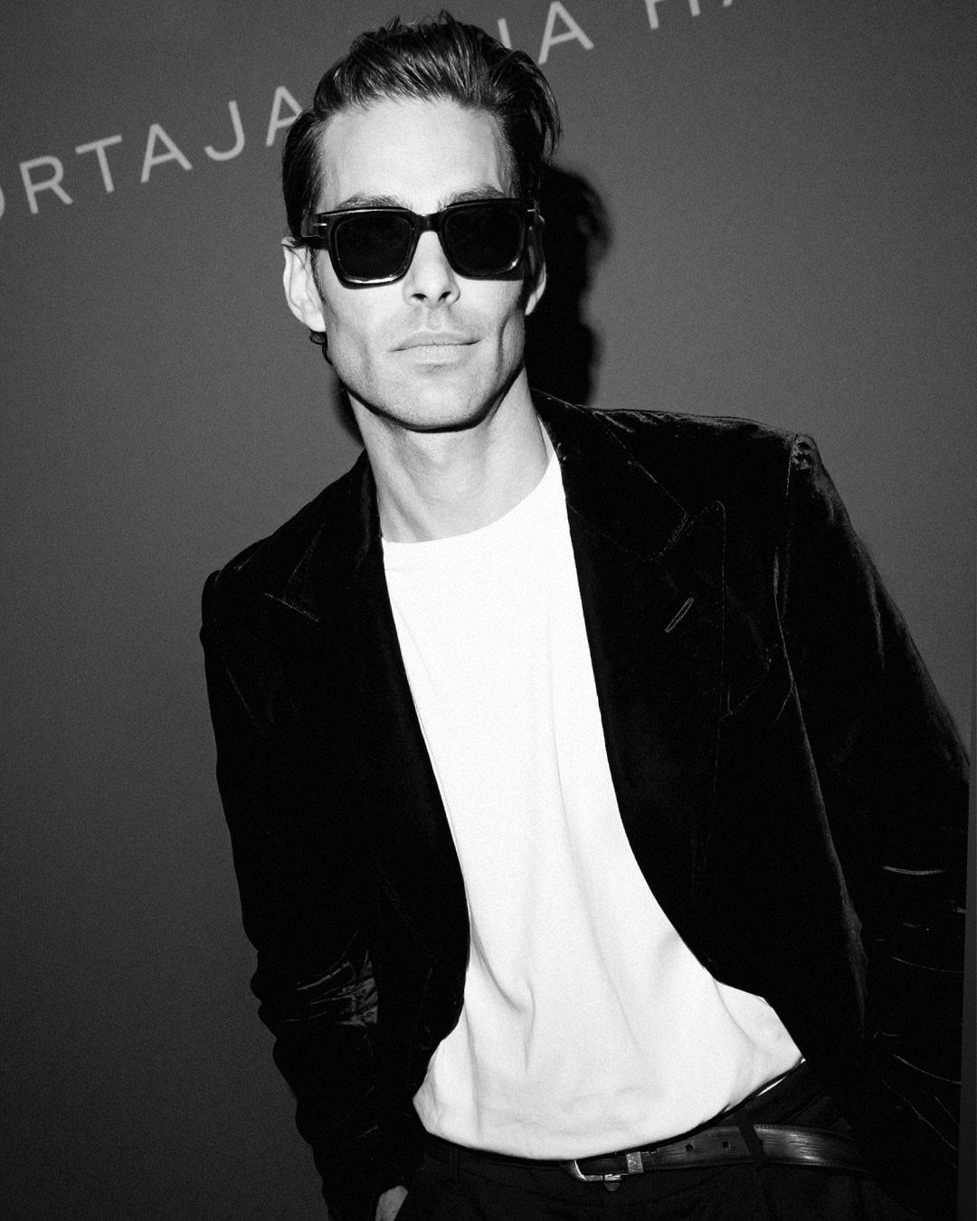 Male model wearing sunglasses, a white t-shirt, and a dark blazer posing against a dark background.