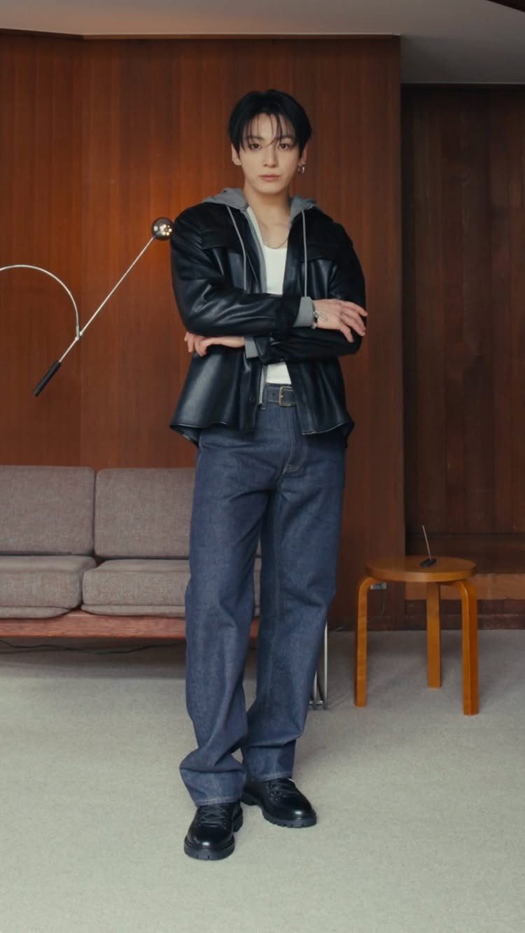 Young man in a leather jacket and denim jeans posing in a modern wood-paneled room.