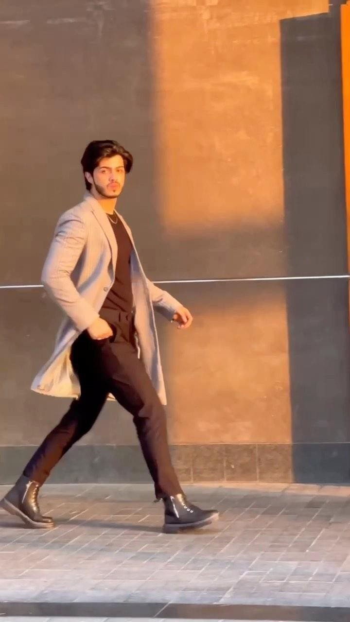 A man in a long grey coat and black trousers walking against a sunlit wall.