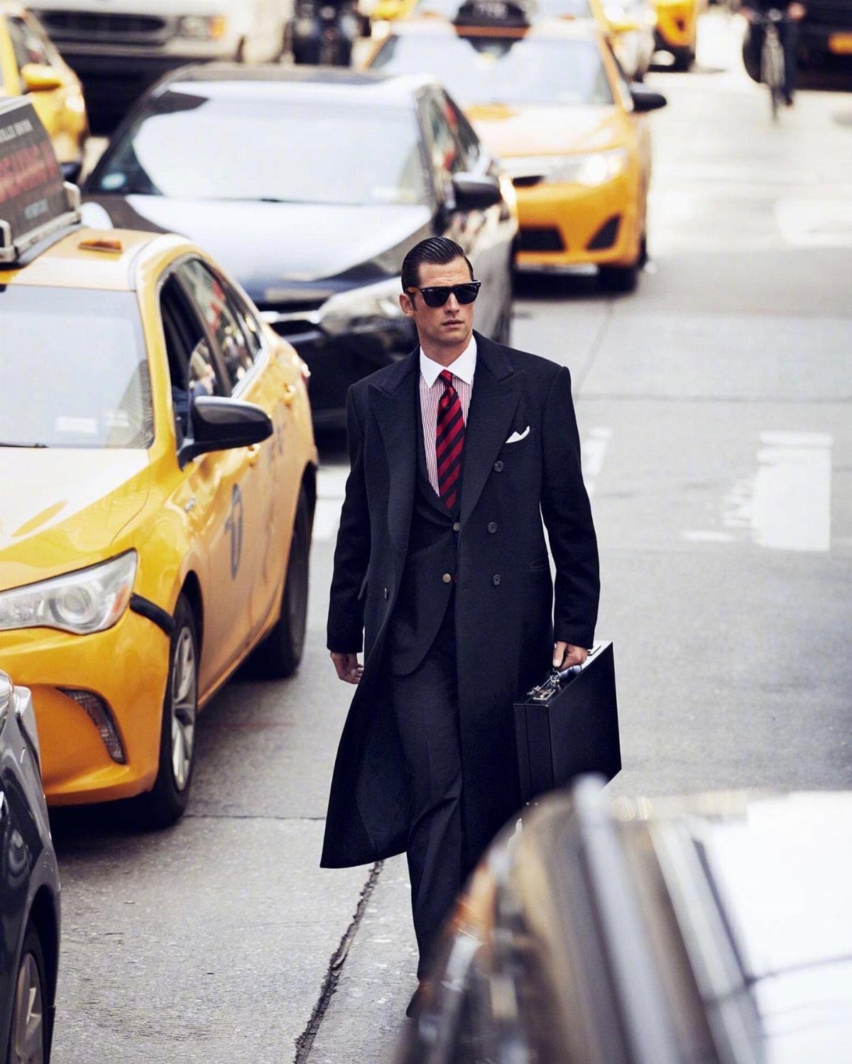 A male model in a black overcoat and sunglasses walking through a city street with yellow taxis.