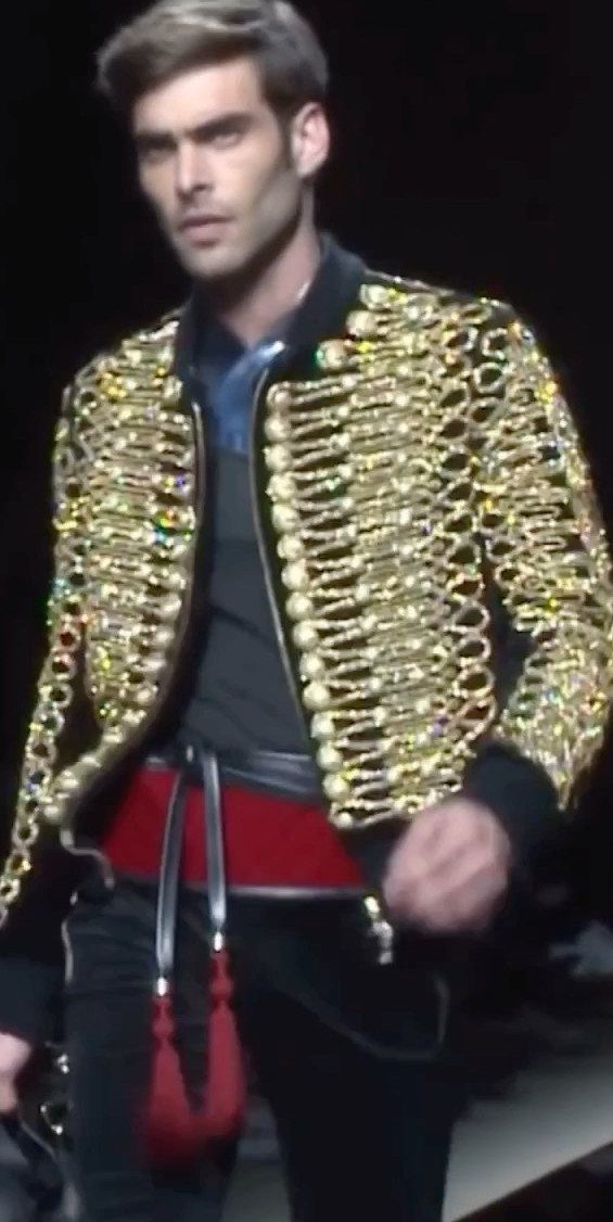 Male model walking a runway in a black jacket decorated with heavy gold chains.