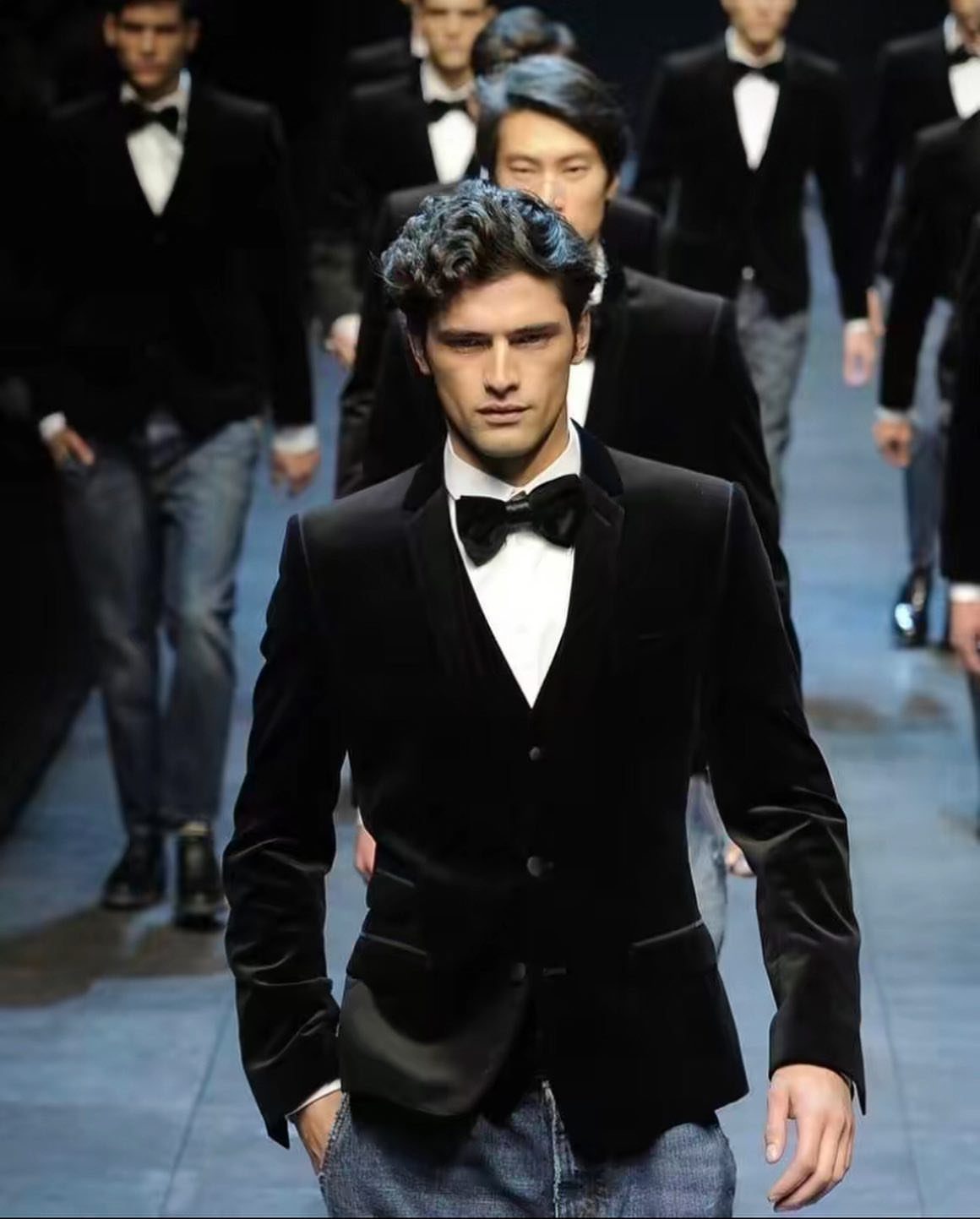 Male model in a black velvet blazer and bowtie walking down a fashion runway.