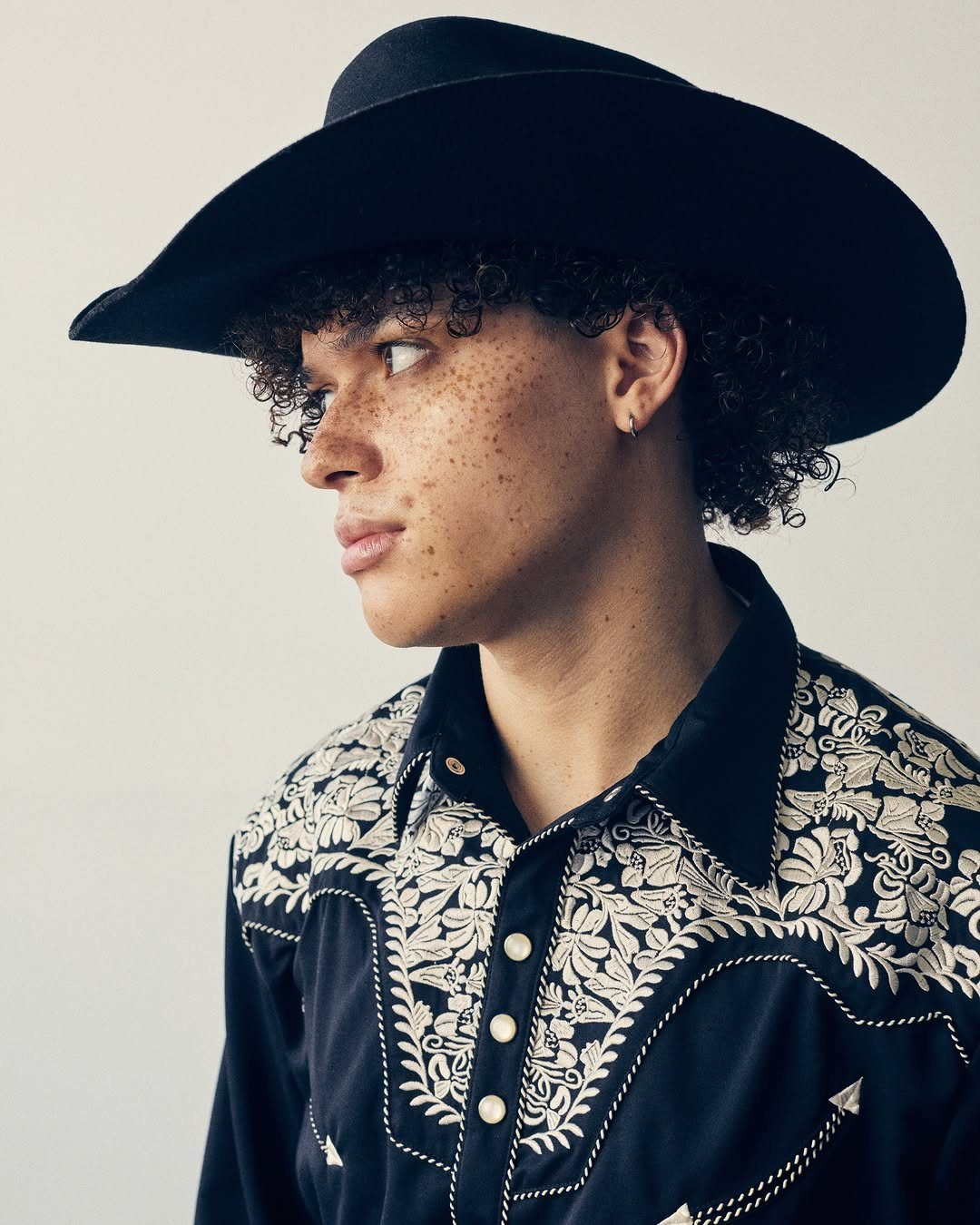 Young man with curly hair and freckles wearing a black cowboy hat and embroidered western shirt.