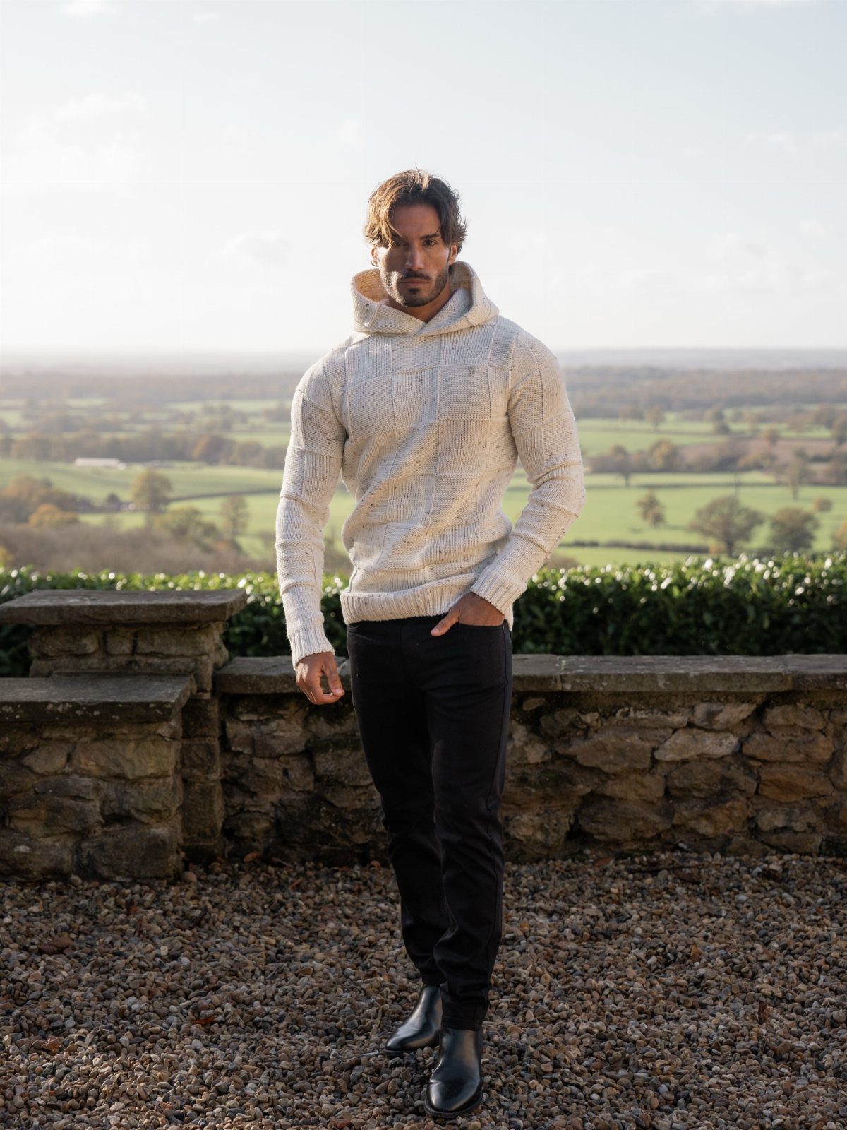 A man wearing a cream cable-knit hooded sweater, black pants, and black boots standing outdoors.