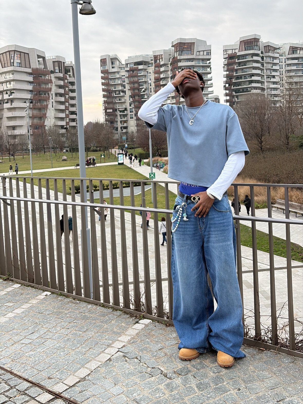 A young man posing in a blue t-shirt, white long-sleeve shirt, and wide-leg denim jeans.