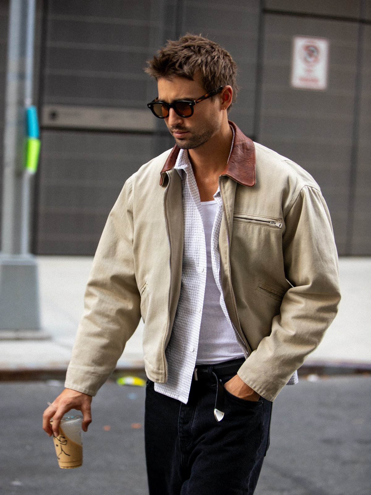 A man in a tan utility jacket and sunglasses walking down a city street.