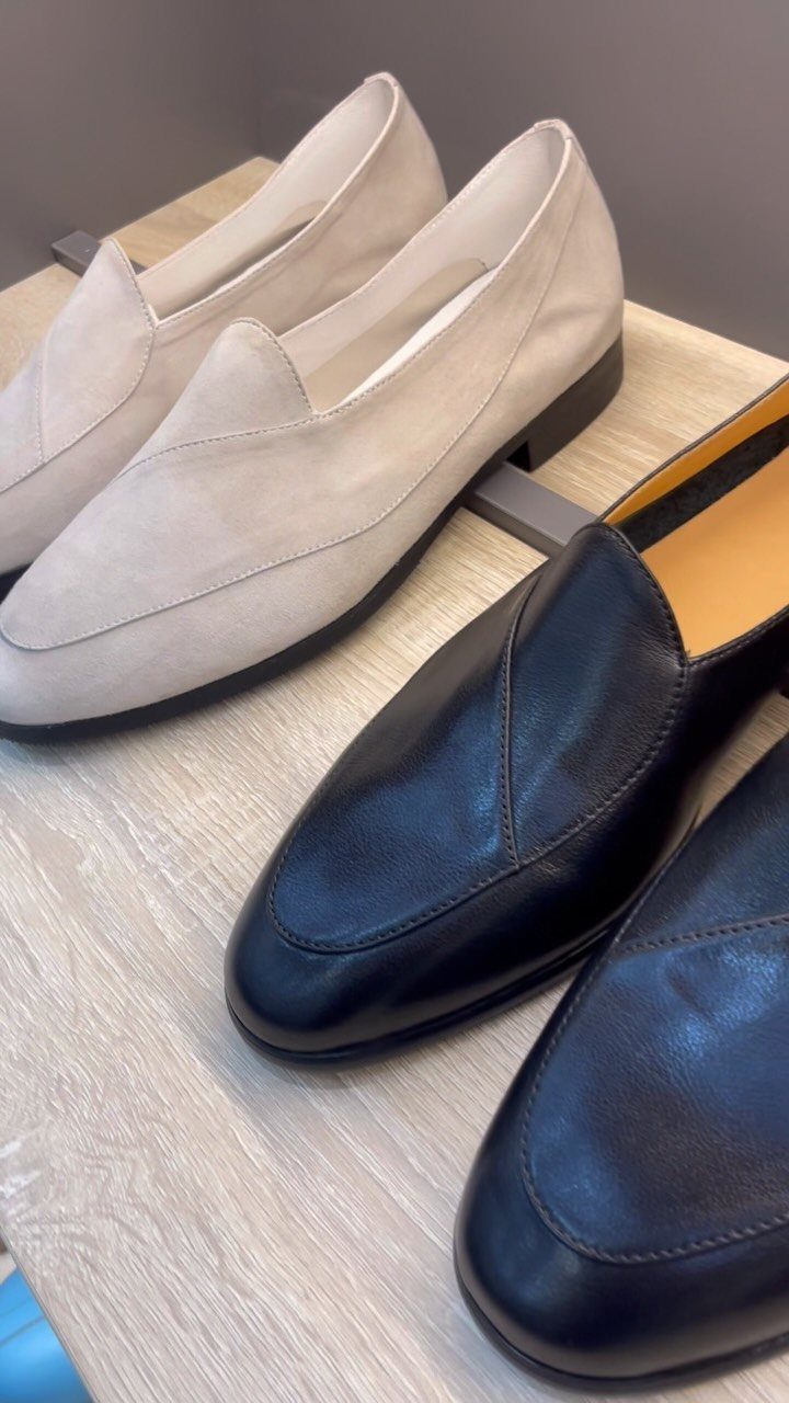Beige suede loafers and black leather loafers resting on a light wooden display shelf.