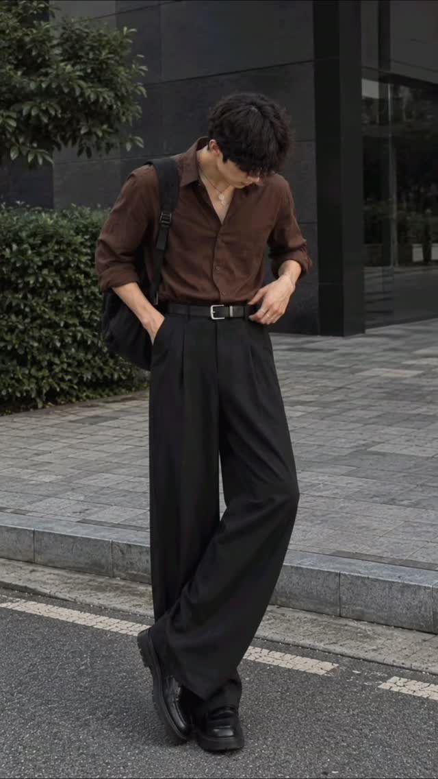 A man in a brown shirt and black trousers posing in black loafers on a city street.