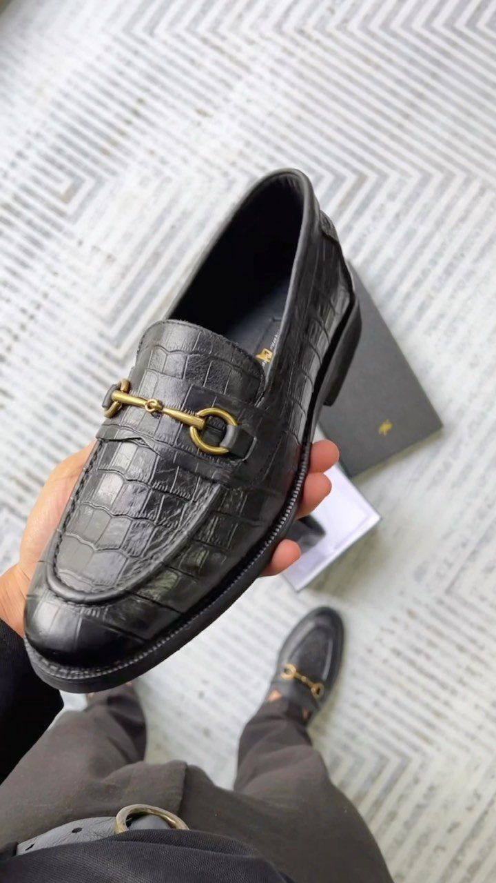 A hand holds a black crocodile-embossed horsebit loafer against a patterned floor.