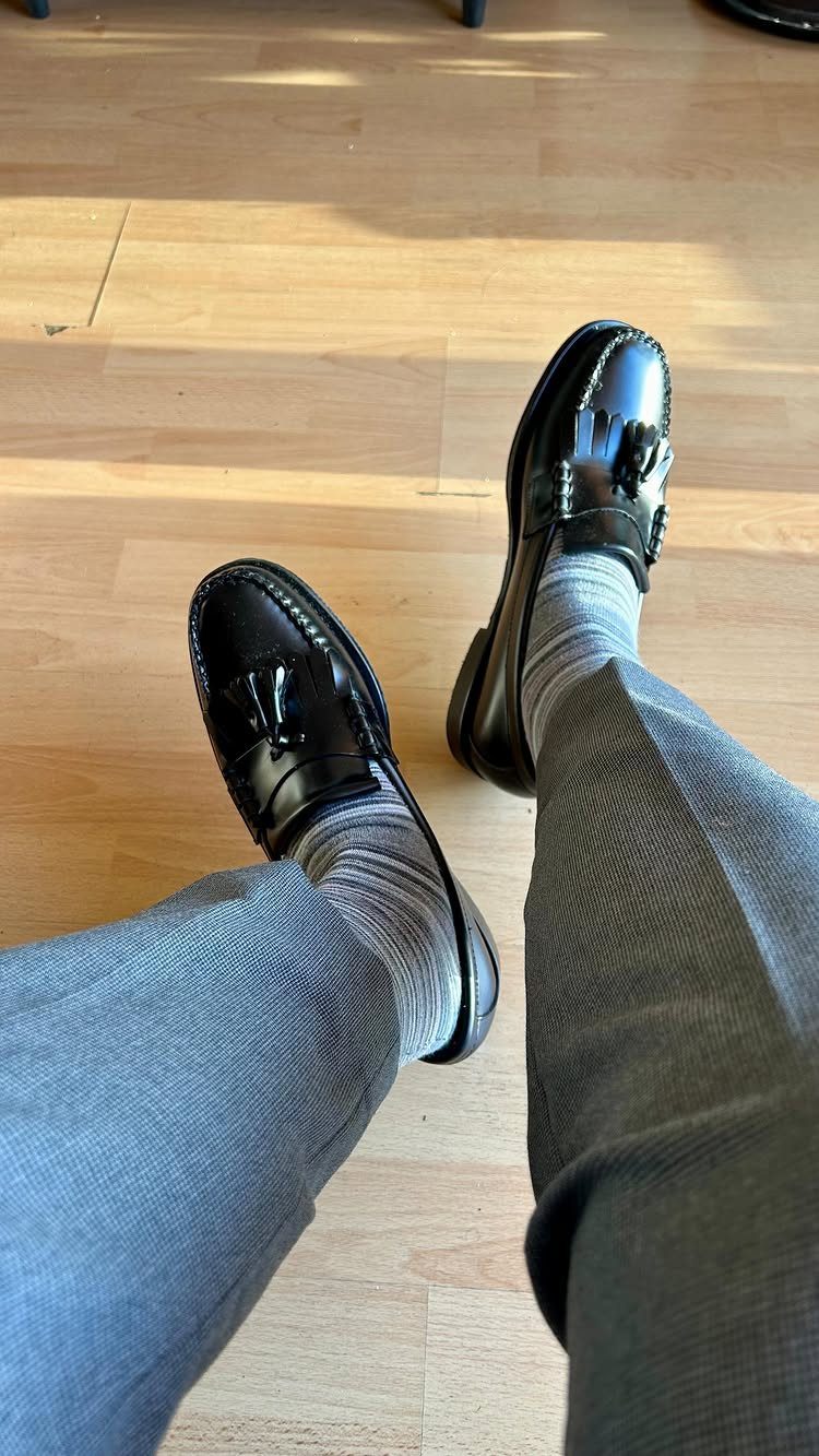 Black leather kiltie loafers with decorative fringes worn with grey socks and trousers.