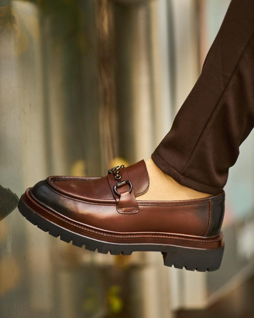 Brown leather loafers with a metal bit detail and thick lugged soles worn with brown trousers.