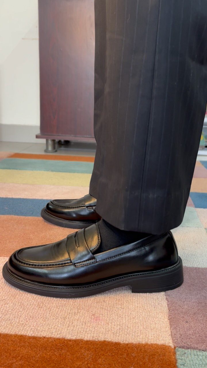 Black leather penny loafers worn with dark pinstripe trousers on a colorful striped rug.