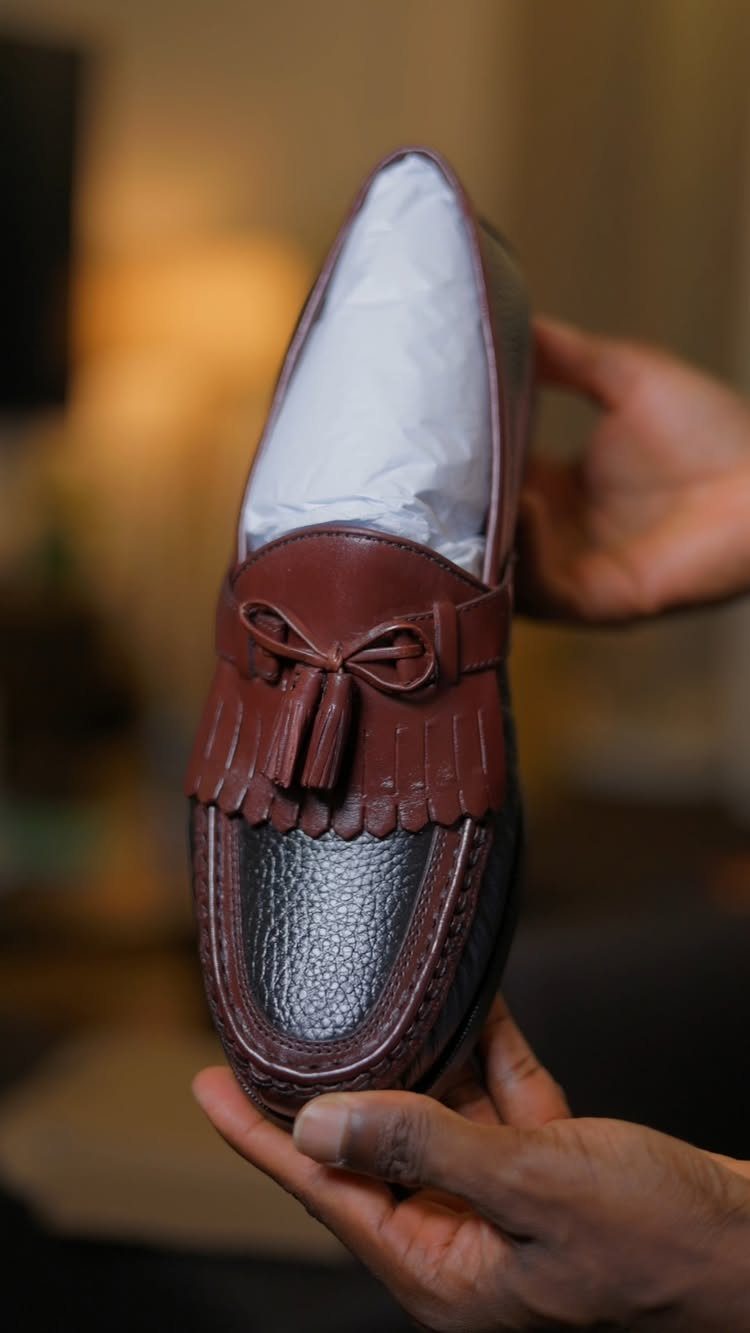 Hands holding a brown leather loafer with decorative tassels and a textured toe cap.