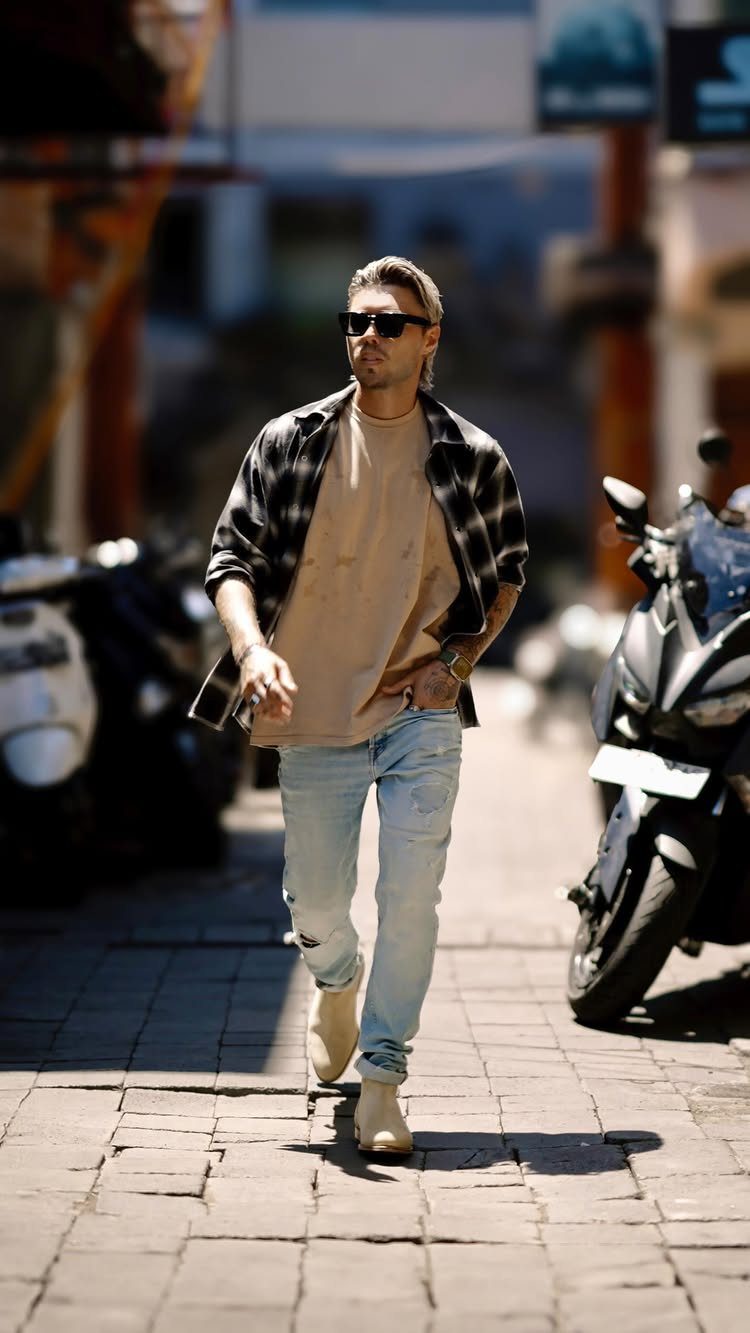 A man walking on a paved street wearing light blue jeans and beige Chelsea boots.