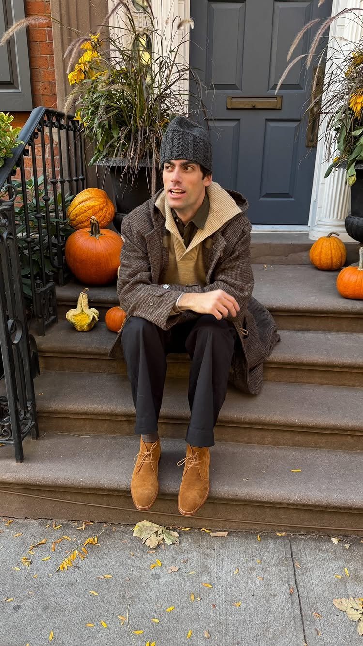 A man in a brown coat and black trousers sits on steps wearing tan suede chukka boots.