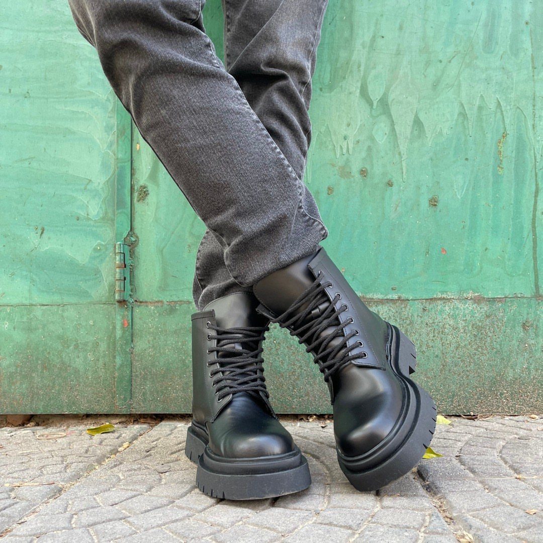 Black leather combat boots with thick soles worn with grey trousers against a green wall.