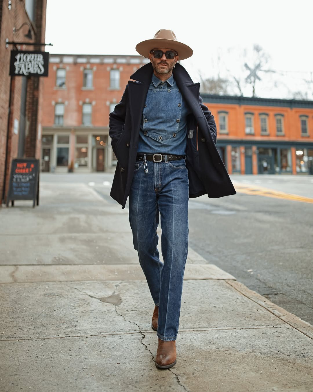 Man in denim outfit and wide-brimmed hat walking down a city street wearing brown boots.
