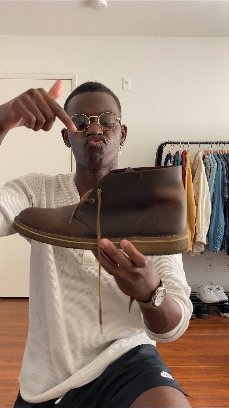 A man holding a brown leather desert boot up to the camera while wearing sunglasses.