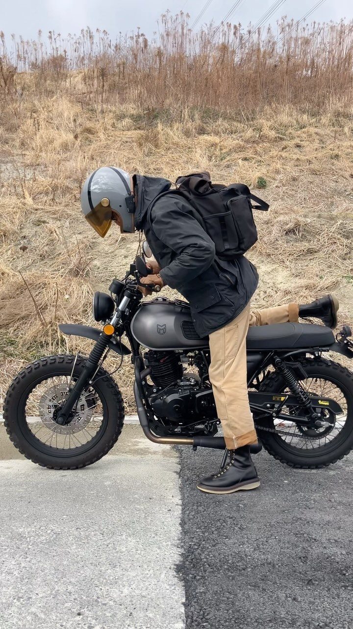 A man in a black jacket and tan pants sits on a grey motorcycle outdoors.