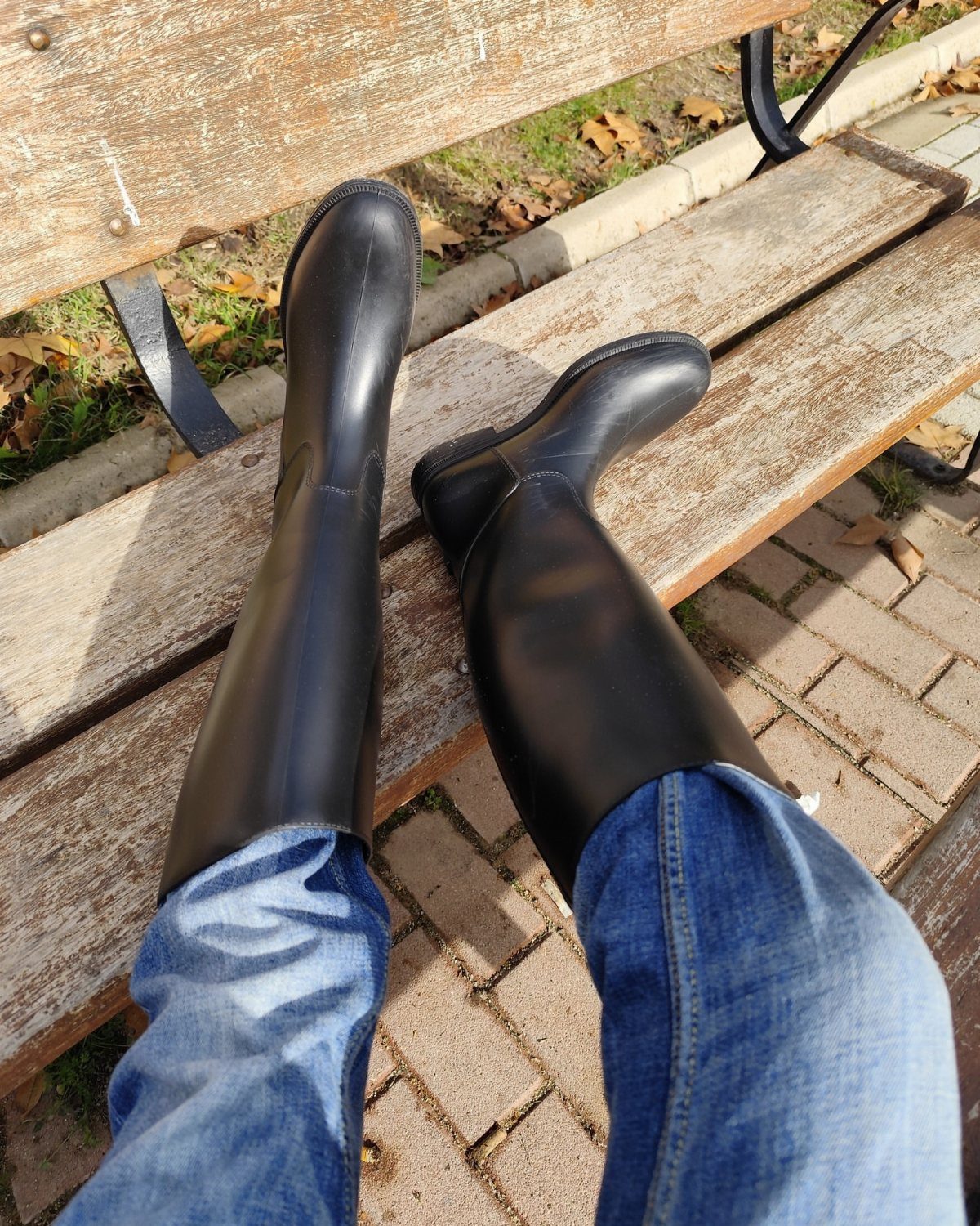 Black rubber boots worn with blue jeans resting on a wooden bench.