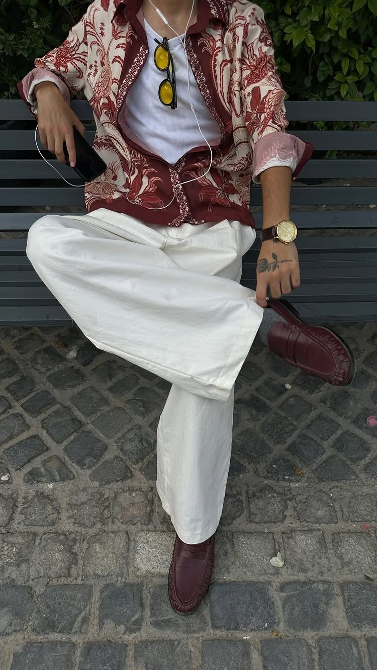 Person wearing a maroon floral patterned shirt, white t-shirt, and white trousers sitting on a bench.