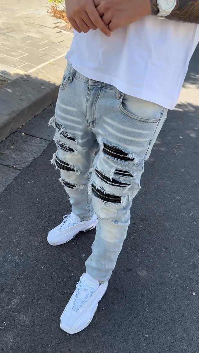 Person wearing light blue distressed jeans with black patches and white sneakers on a sidewalk.