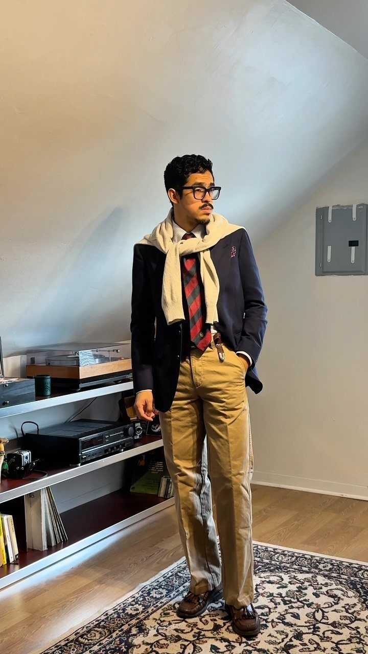 Man in navy blazer, striped tie, tan trousers, and a sweater draped over his shoulders.