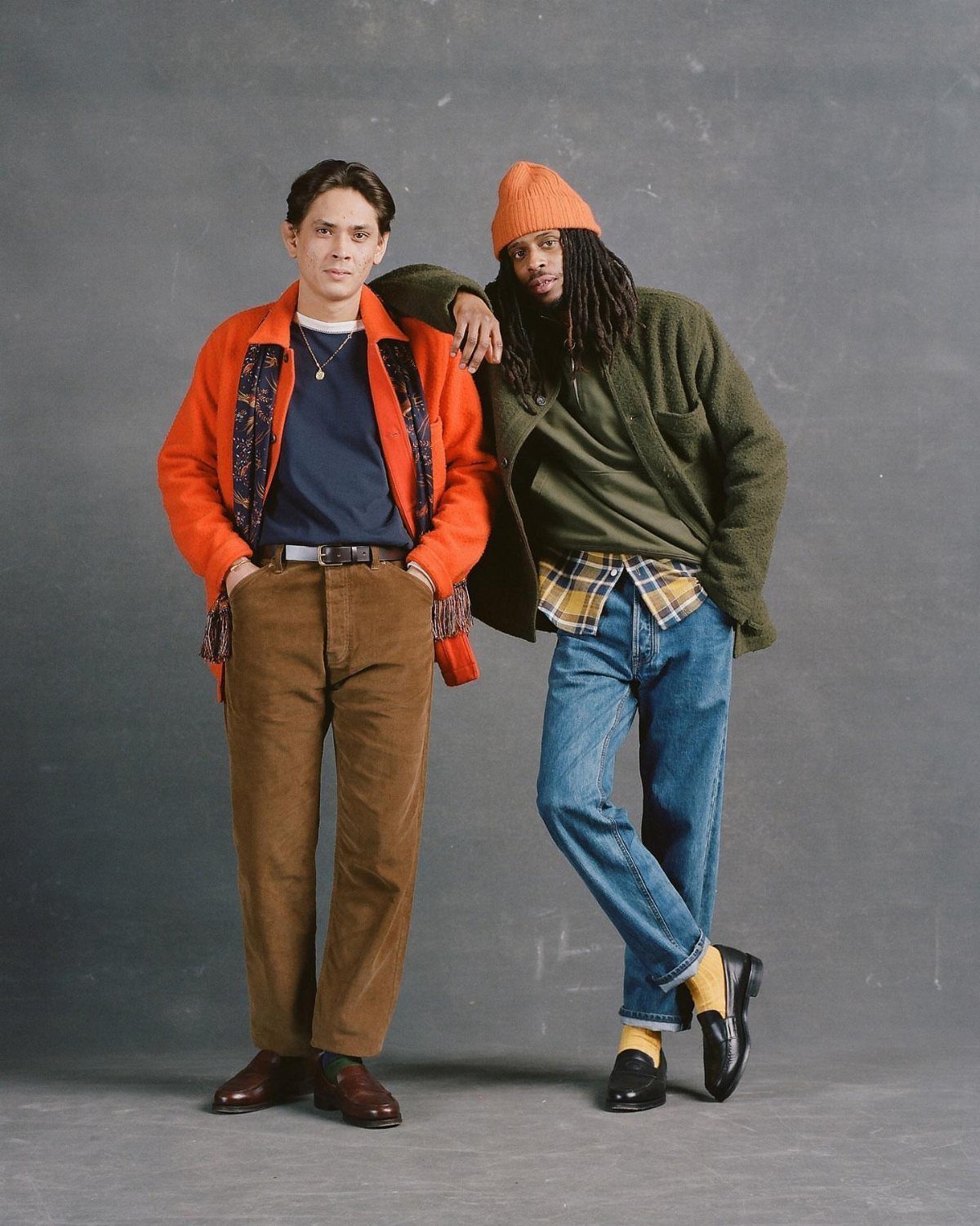 Two men posing in colorful textured jackets, plaid shirts, and denim jeans against a grey backdrop.