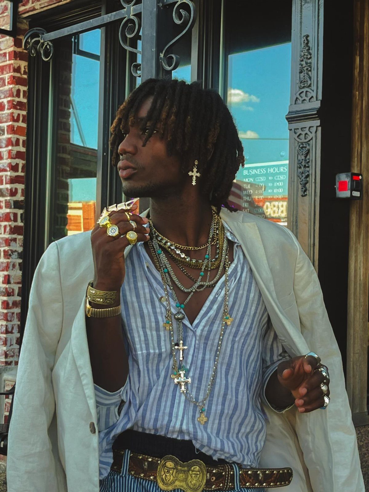 Man with dreadlocks wearing a white blazer, striped shirt, heavy gold jewelry, and an ornate gold belt.