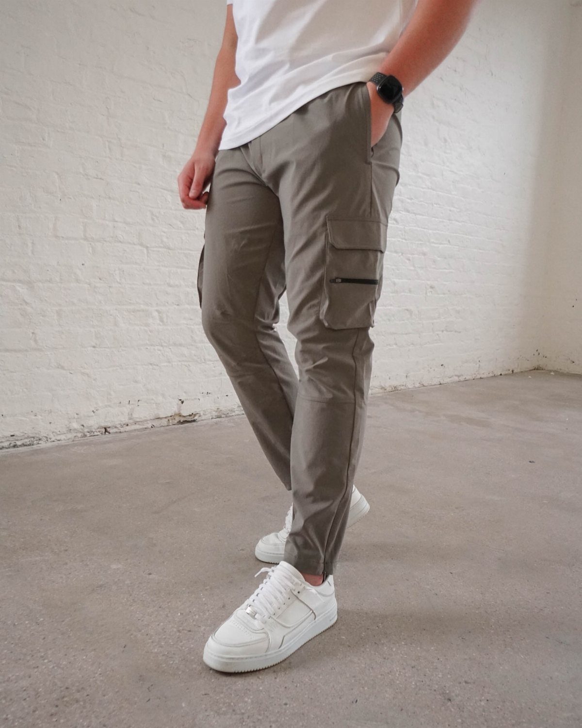 Man wearing grey cargo pants and white sneakers standing against a white brick wall.