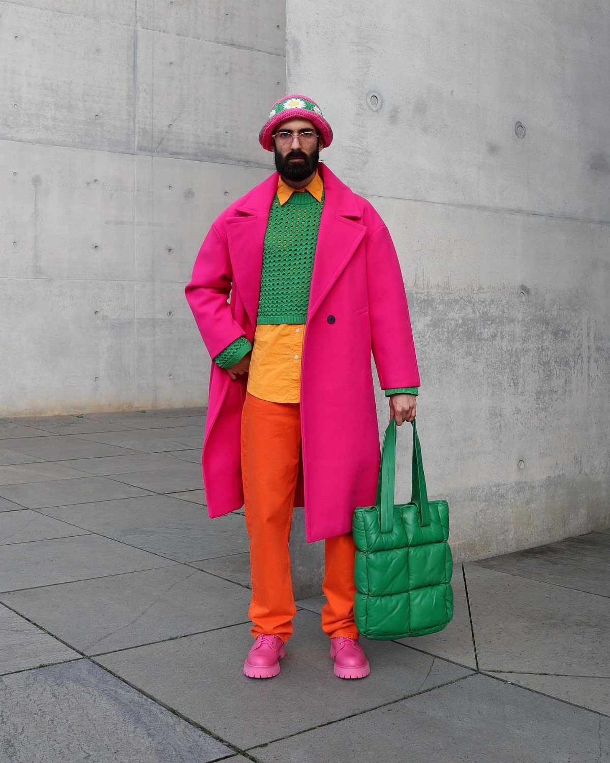 Man in vibrant pink coat, green sweater, and orange trousers standing against a concrete wall.