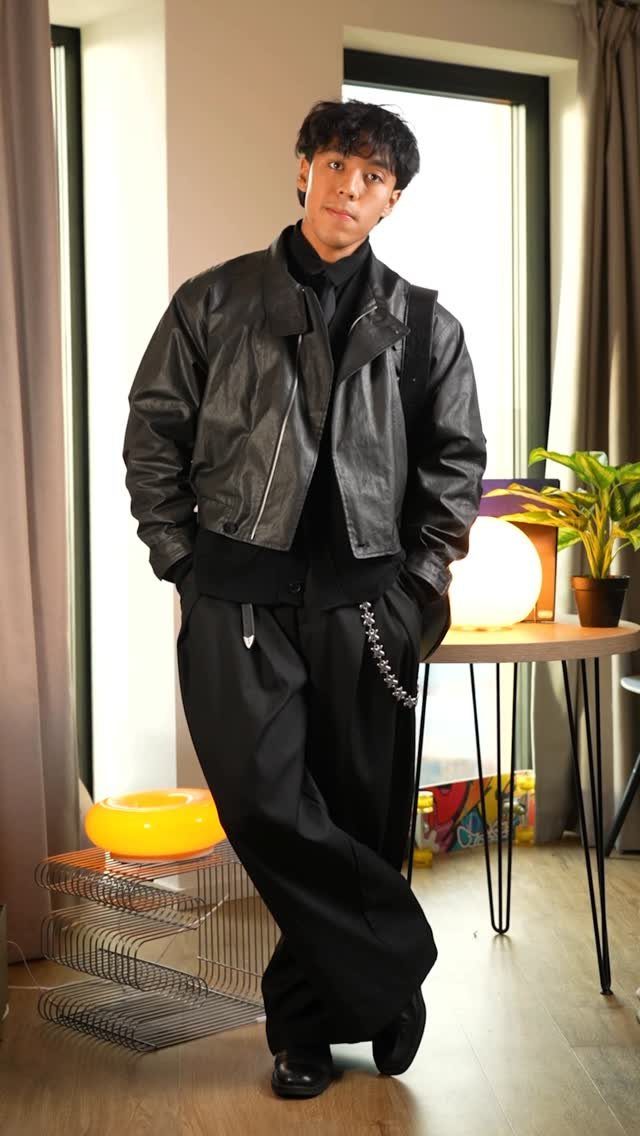 Man wearing a black leather cropped jacket and wide-leg black trousers standing indoors.
