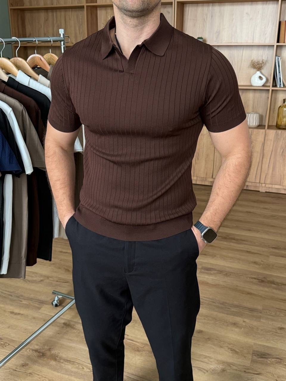 Man wearing a brown ribbed polo shirt and black trousers standing in a clothing store.