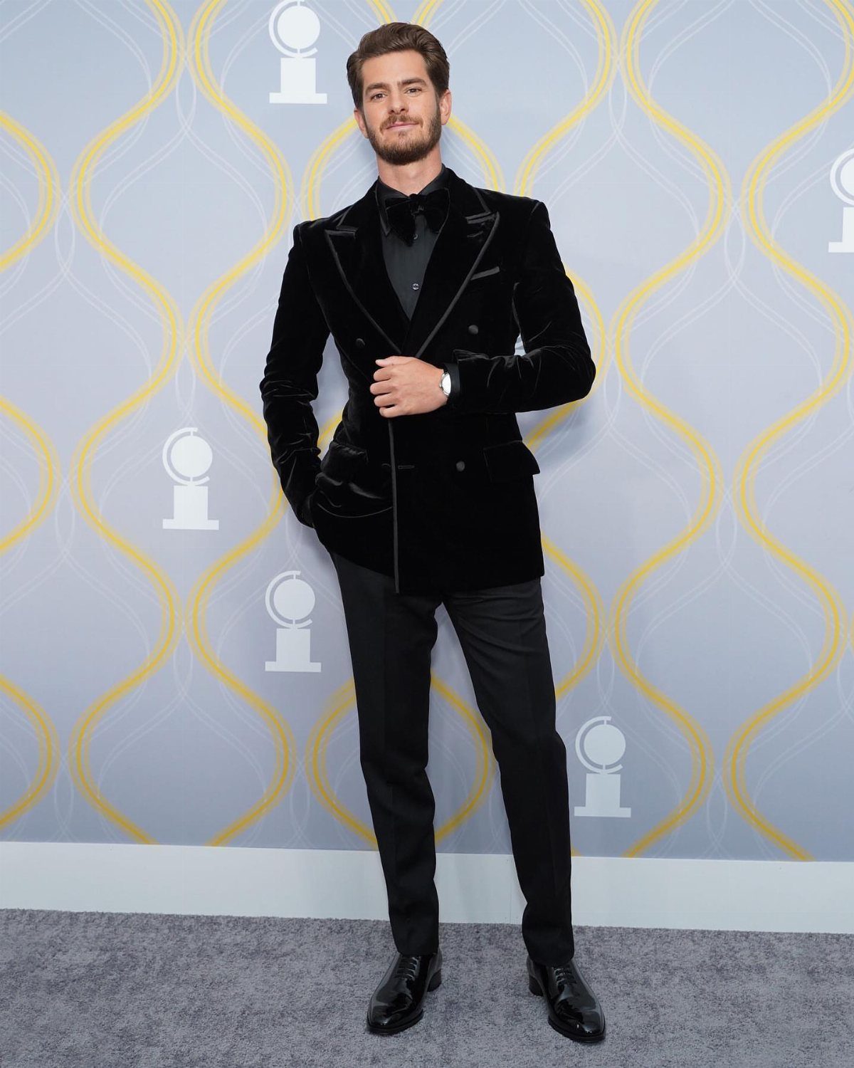 Man wearing a black velvet blazer and bow tie standing against a patterned backdrop.