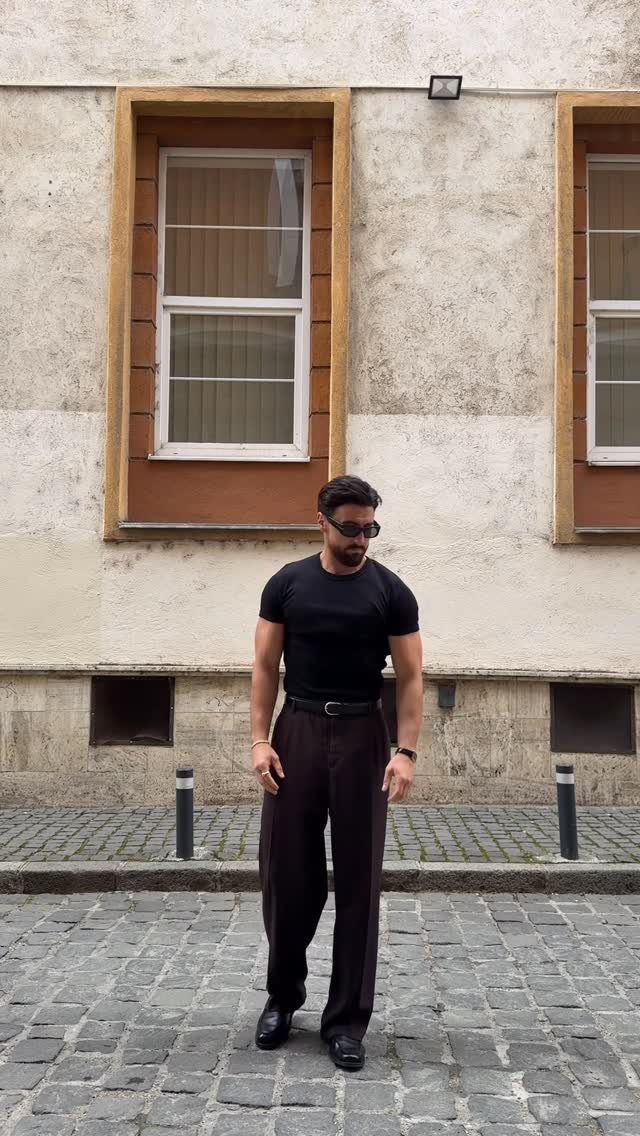 Man in black t-shirt and dark brown wide-leg trousers standing on a cobblestone street.