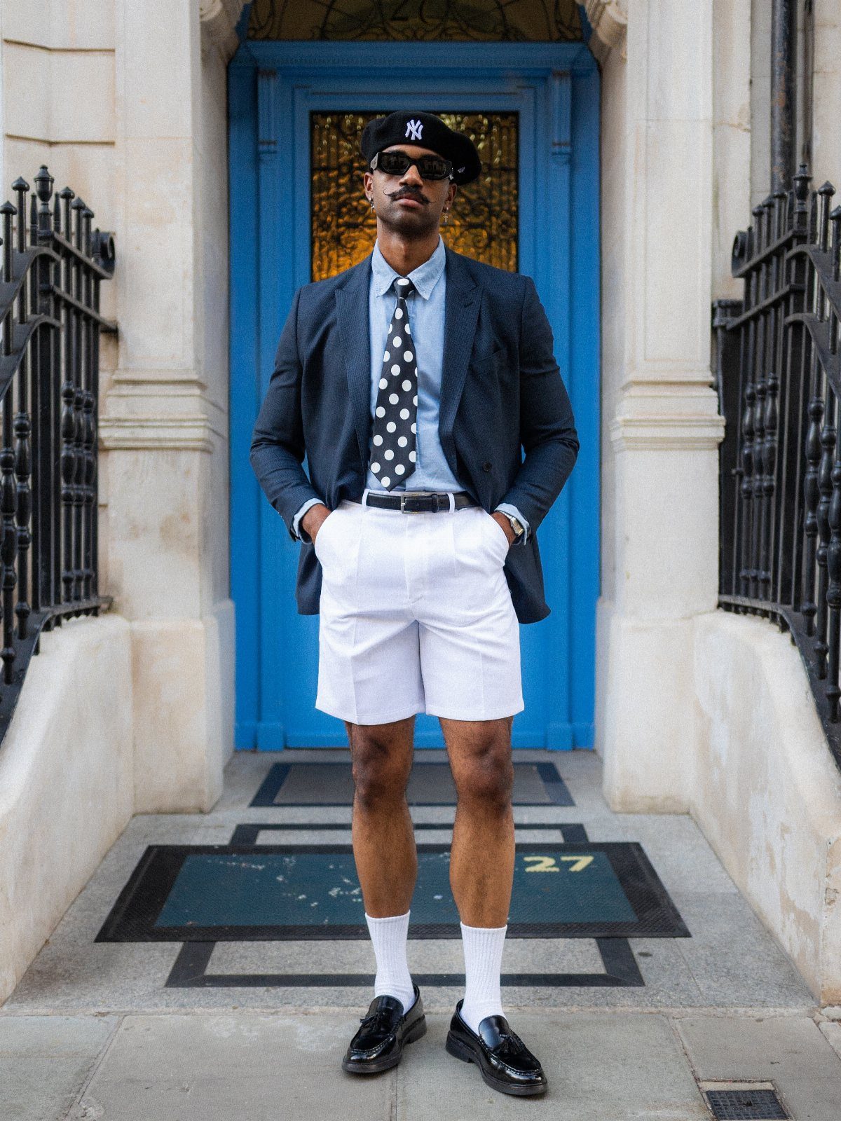 Man in a dark blazer, polka dot tie, and white shorts standing before a blue door.