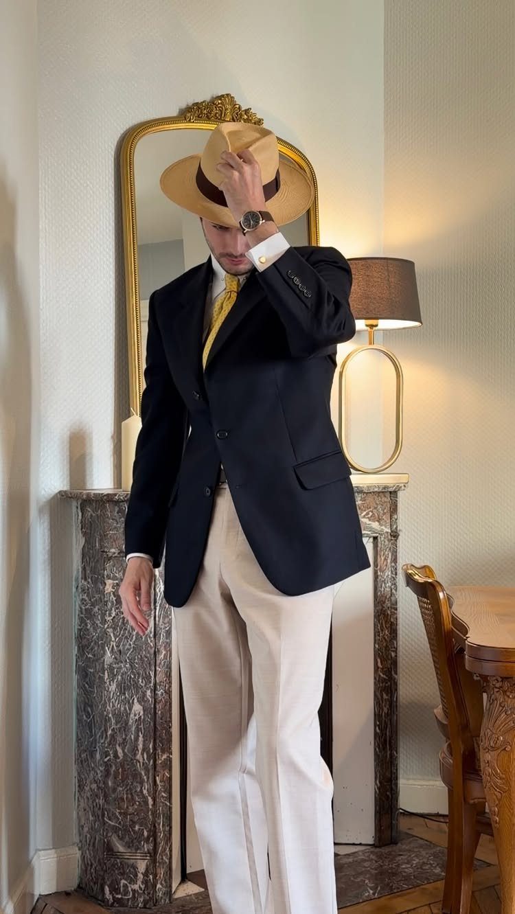 Man in a navy blazer, yellow tie, cream trousers, and a tan fedora hat.