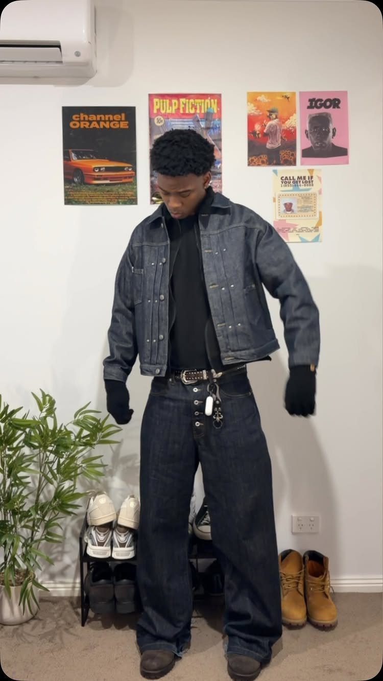 A man wearing a dark denim jacket, black shirt, and wide-leg jeans standing in a room.