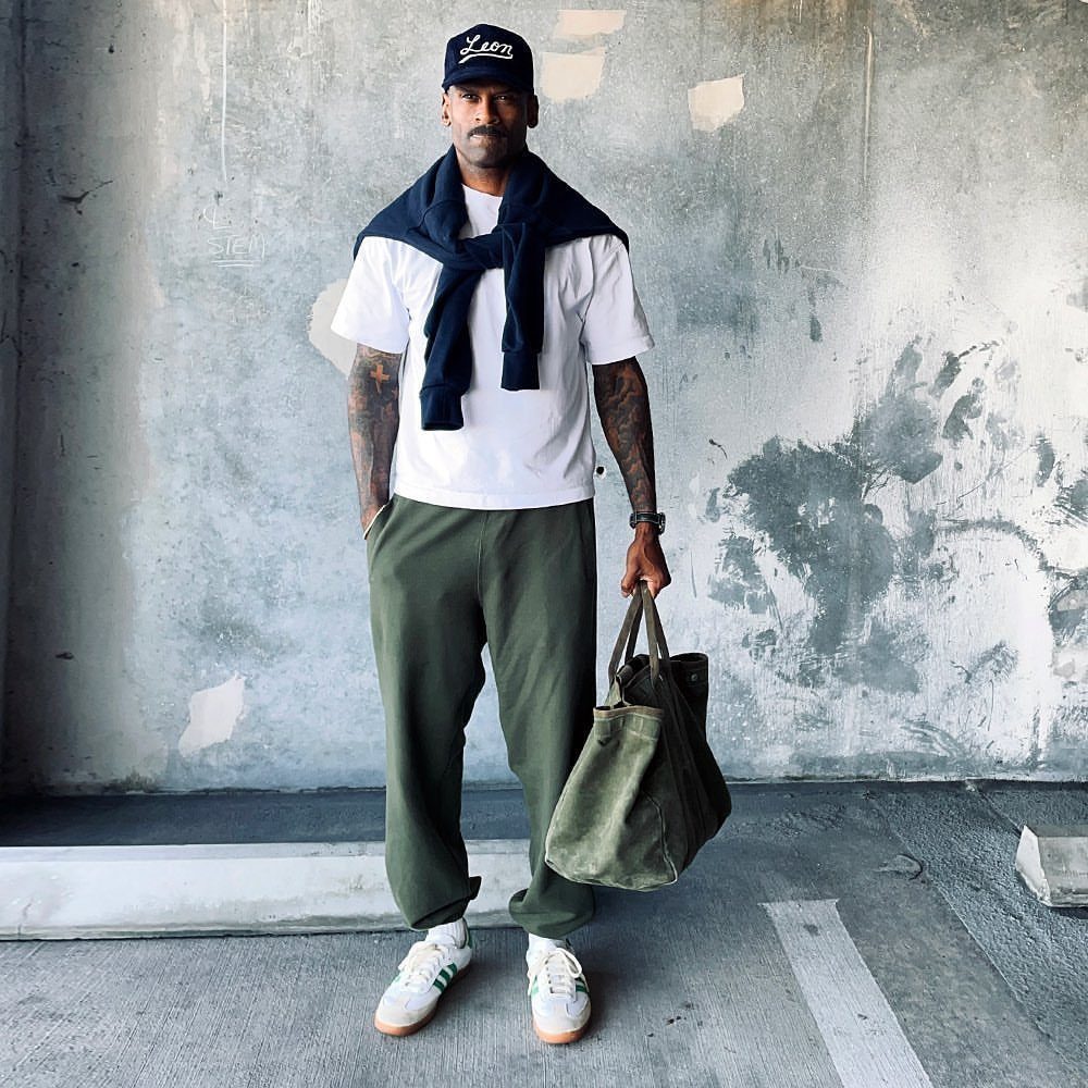 Man in white t-shirt, olive trousers, and white sneakers standing against a concrete wall.