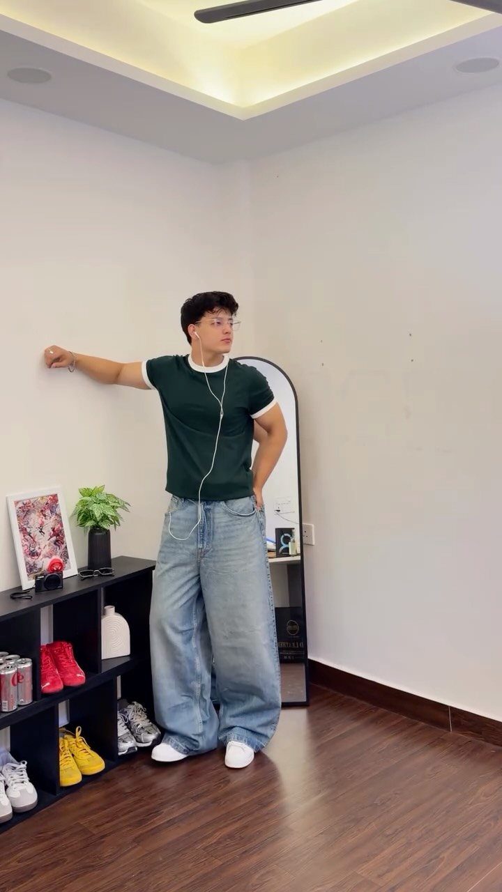 A young man posing in a dark green t-shirt and oversized light blue denim jeans.